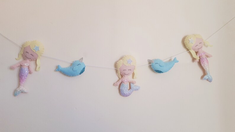 May include: A felt garland with three mermaids and two narwhals. The mermaids have blonde hair and pink and purple tails. The narwhals are blue. The garland is strung on a white string.