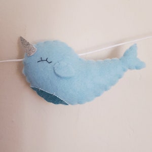 May include: A light blue felt narwhal with a silver glitter horn and black eyes. The narwhal is hanging from a white string.