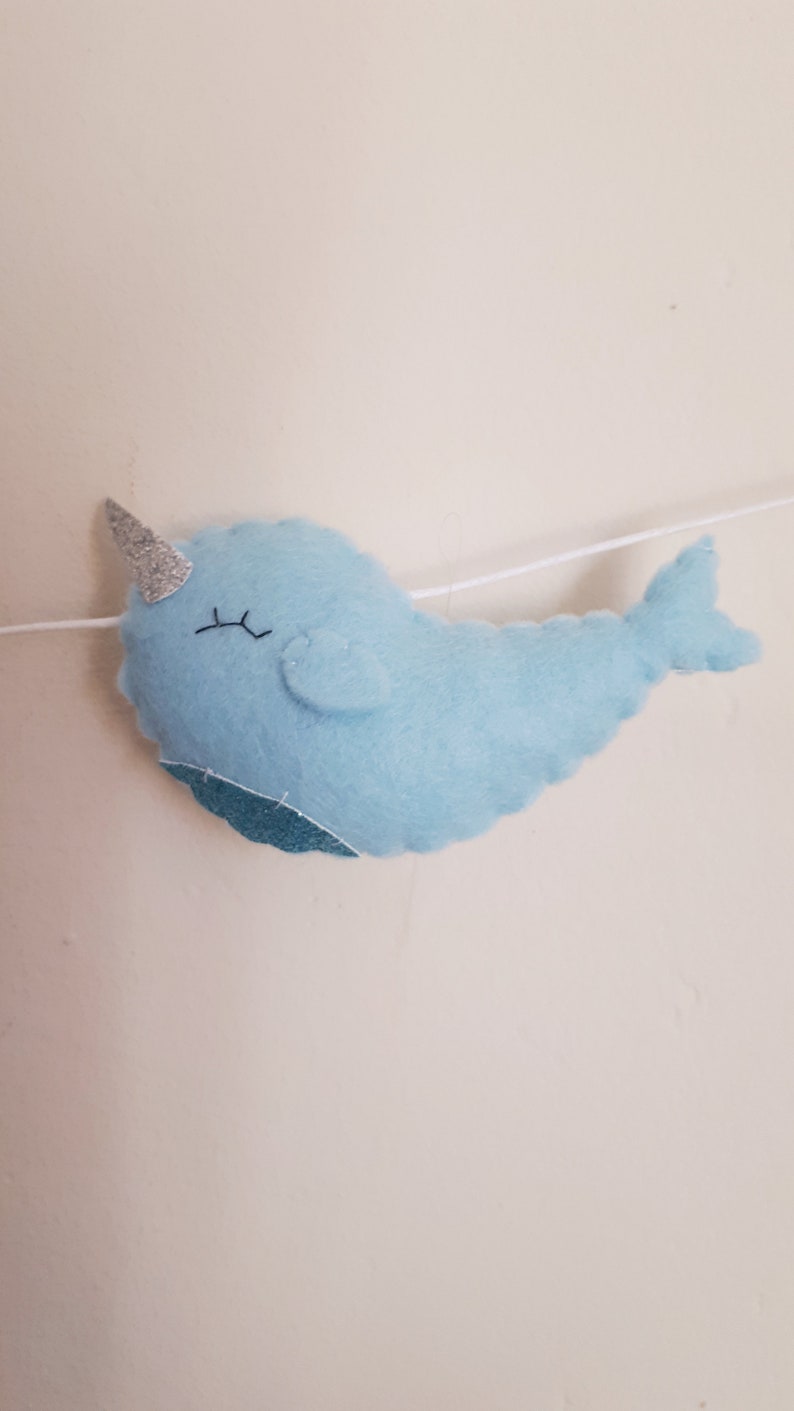 May include: A light blue felt narwhal with a silver glitter horn and black eyes. The narwhal is hanging from a white string.