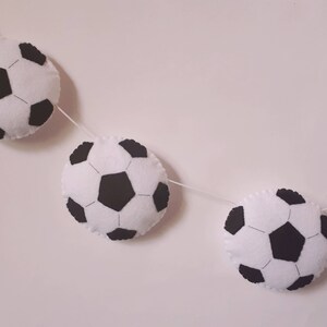 Puede incluir: Una guirnalda de bolas de f&uacute;tbol de fieltro blanco y negro con tres bolas colgando de una cuerda blanca.