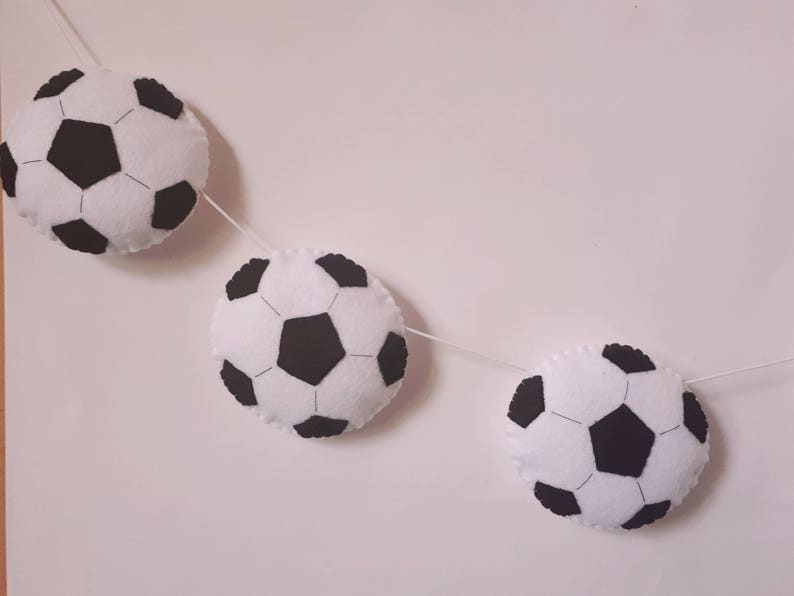 Puede incluir: Una guirnalda de bolas de f&uacute;tbol de fieltro blanco y negro con tres bolas colgando de una cuerda blanca.