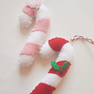 May include: Two handmade candy cane ornaments. One has pink stripes and a pink glitter bow with a silver bell. The other has red stripes and a red bow with green holly leaves. Both are white and made of felt.