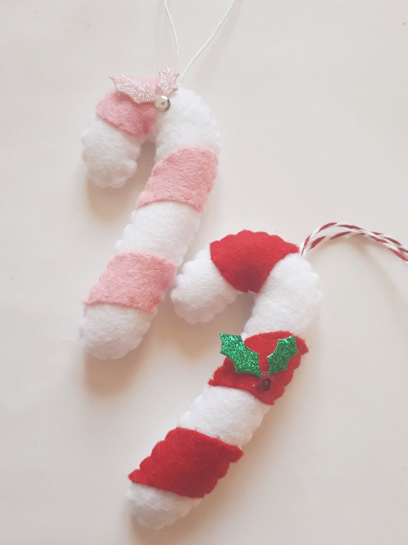 May include: Two handmade candy cane ornaments. One has pink stripes and a pink glitter bow with a silver bell. The other has red stripes and a red bow with green holly leaves. Both are white and made of felt.