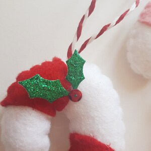 May include: A felt Christmas ornament shaped like a candy cane, with red and white stripes. A green glitter holly leaf and a red bead are attached. The ornament hangs from a red and white twisted cord. Festive holiday decor.
