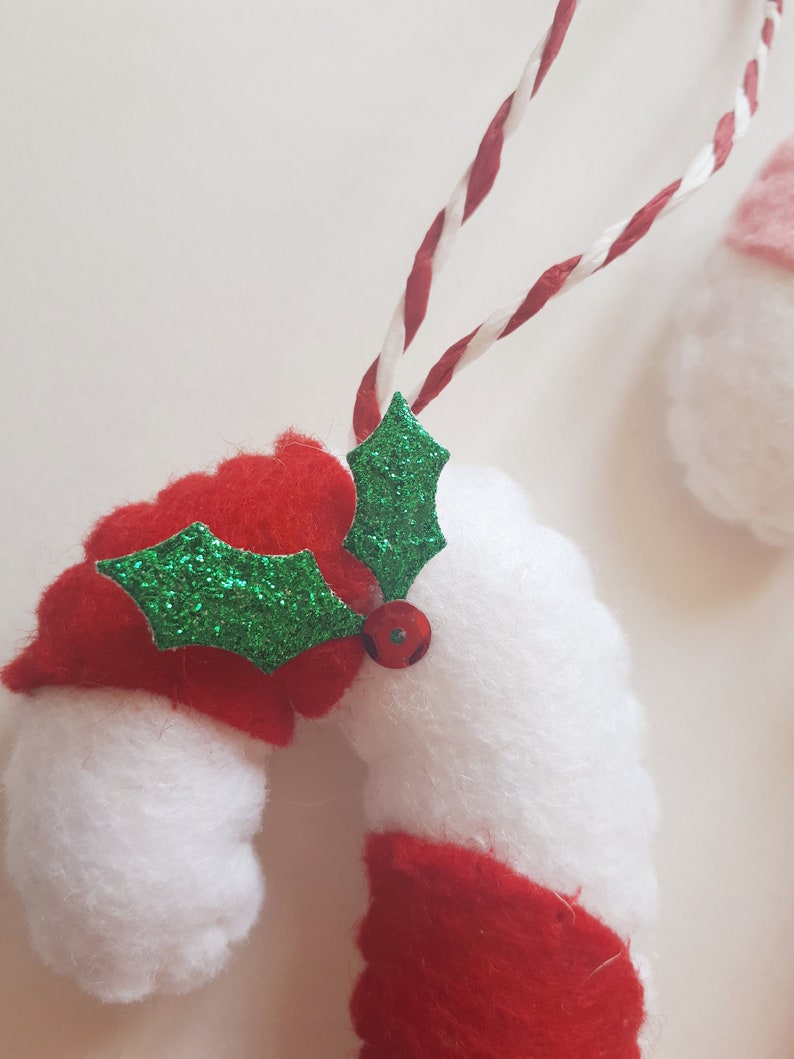 May include: A felt Christmas ornament shaped like a candy cane, with red and white stripes. A green glitter holly leaf and a red bead are attached. The ornament hangs from a red and white twisted cord. Festive holiday decor.