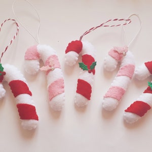 May include: Five handmade candy cane ornaments. Each candy cane is white with red and pink stripes, and some have decorative holly or glittery accents. They are strung with white and red twine for hanging.