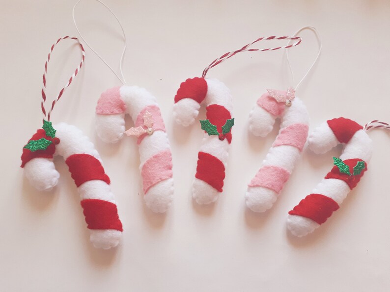 May include: Five handmade candy cane ornaments. Each candy cane is white with red and pink stripes, and some have decorative holly or glittery accents. They are strung with white and red twine for hanging.