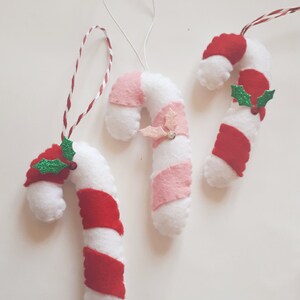 May include: Three handmade Christmas candy cane ornaments. The candy canes are white with red or pink stripes, and some have holly accents. Each ornament has a string for hanging.