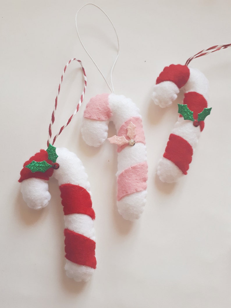 May include: Three handmade Christmas candy cane ornaments. The candy canes are white with red or pink stripes, and some have holly accents. Each ornament has a string for hanging.