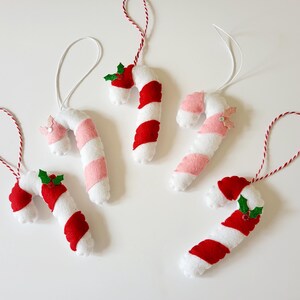 May include: A collection of handmade candy cane ornaments. Each ornament is white with red and pink stripes, and some feature holly accents. They have white or red and white twine for hanging, perfect for holiday decor.