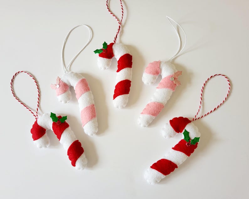 May include: A collection of handmade candy cane ornaments. Each ornament is white with red and pink stripes, and some feature holly accents. They have white or red and white twine for hanging, perfect for holiday decor.
