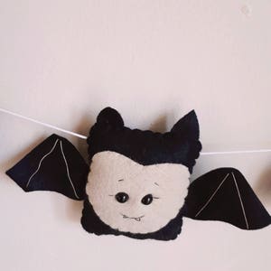 May include: A felt bat decoration with a light beige face, black wings, and a black head. The bat has two black button eyes and a stitched smile. The wings are attached to a white string, suggesting a garland or banner.