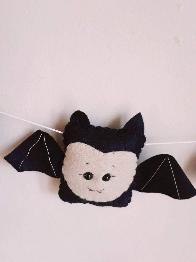 May include: A felt bat decoration with a light beige face, black wings, and a black head. The bat has two black button eyes and a stitched smile. The wings are attached to a white string, suggesting a garland or banner.