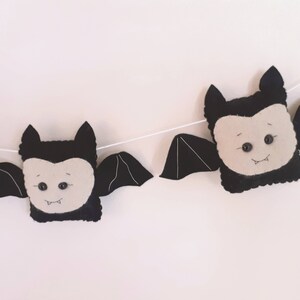 May include: A Halloween garland with felt bat decorations. Each bat has a black body, cream face, and black wings. The bats are strung on a white string, creating a festive banner for Halloween decorations.