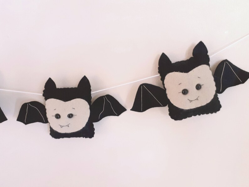 May include: A Halloween garland with felt bat decorations. Each bat has a black body, cream face, and black wings. The bats are strung on a white string, creating a festive banner for Halloween decorations.