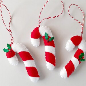 May include: Three handmade Christmas ornaments shaped like candy canes. Each candy cane is white with red stripes and a small green holly accent. They hang from red and white twine.