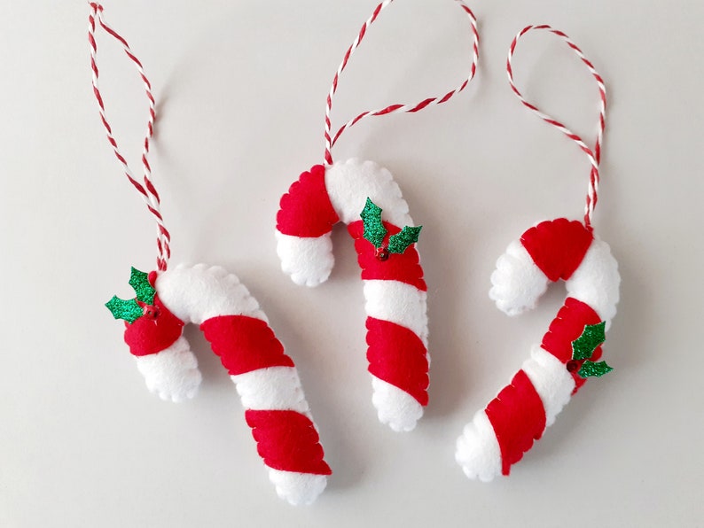 May include: Three handmade Christmas ornaments shaped like candy canes. Each candy cane is white with red stripes and a small green holly accent. They hang from red and white twine.