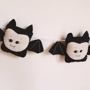 May include: A Halloween garland with plush bat decorations. Each bat has a black body, wings, and ears, with a light beige face and small black button eyes. The bats are strung together on a white string.