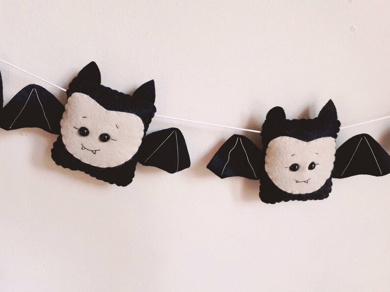 May include: A Halloween garland with plush bat decorations. Each bat has a black body, wings, and ears, with a light beige face and small black button eyes. The bats are strung together on a white string.
