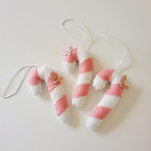 May include: Three handmade candy cane ornaments. Each ornament is white with pink stripes and a small pink bow. The ornaments are made of felt and have white string loops for hanging. These festive decorations are perfect for a Christmas tree.