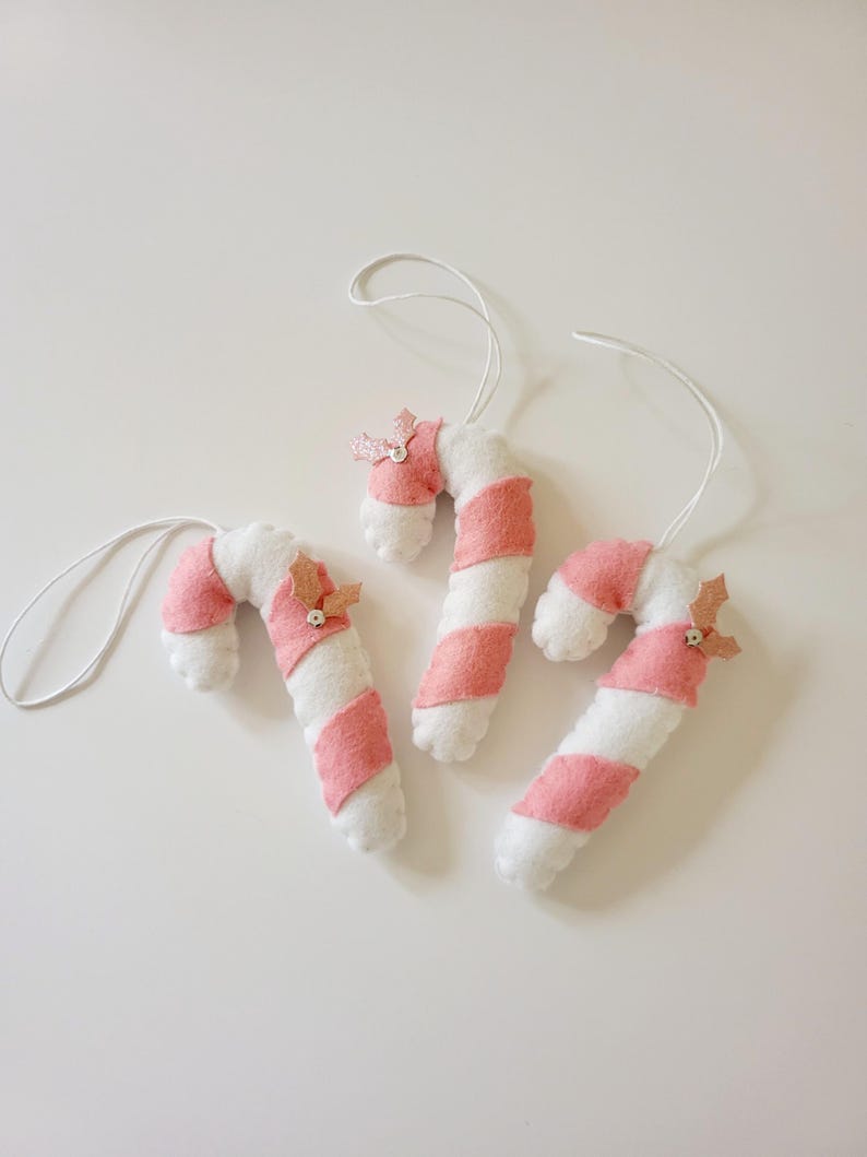 May include: Three handmade candy cane ornaments. Each ornament is white with pink stripes and a small pink bow. The ornaments are made of felt and have white string loops for hanging. These festive decorations are perfect for a Christmas tree.