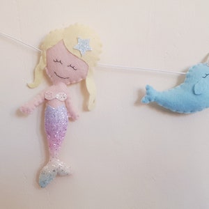 May include: A felt garland with a mermaid and a narwhal. The mermaid is pink with a glittery purple tail and a yellow star on her head. The narwhal is blue with a silver horn.