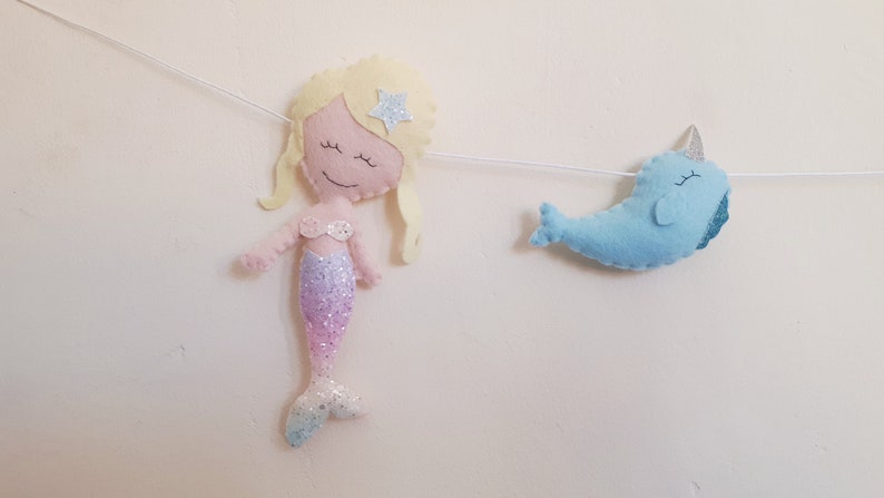 May include: A felt garland with a mermaid and a narwhal. The mermaid is pink with a glittery purple tail and a yellow star on her head. The narwhal is blue with a silver horn.