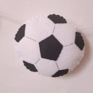 Puede incluir: Un bal&oacute;n de f&uacute;tbol de fieltro blanco con costuras negras y paneles de fieltro negro. El bal&oacute;n est&aacute; colgado de una cuerda blanca.