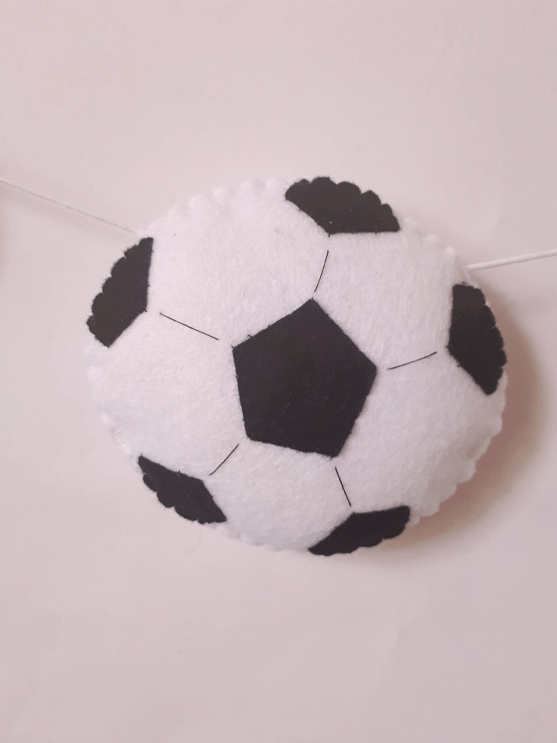 Puede incluir: Un bal&oacute;n de f&uacute;tbol de fieltro blanco con costuras negras y paneles de fieltro negro. El bal&oacute;n est&aacute; colgado de una cuerda blanca.