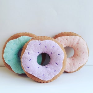 Puede incluir: Tres donuts de fieltro en colores pastel. Los donuts son azul claro, lavanda y rosa. Cada donut tiene un anillo de fieltro marrón y está decorado con pequeñas chispas blancas de fieltro.