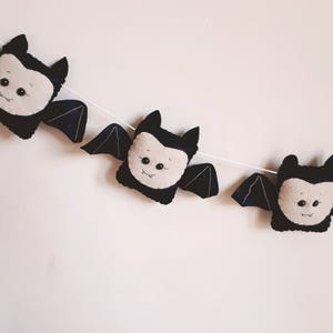 May include: A decorative garland featuring three plush bats. Each bat has black wings and body, with a cream-coloured face. The bats are strung on a white string, ideal for Halloween.