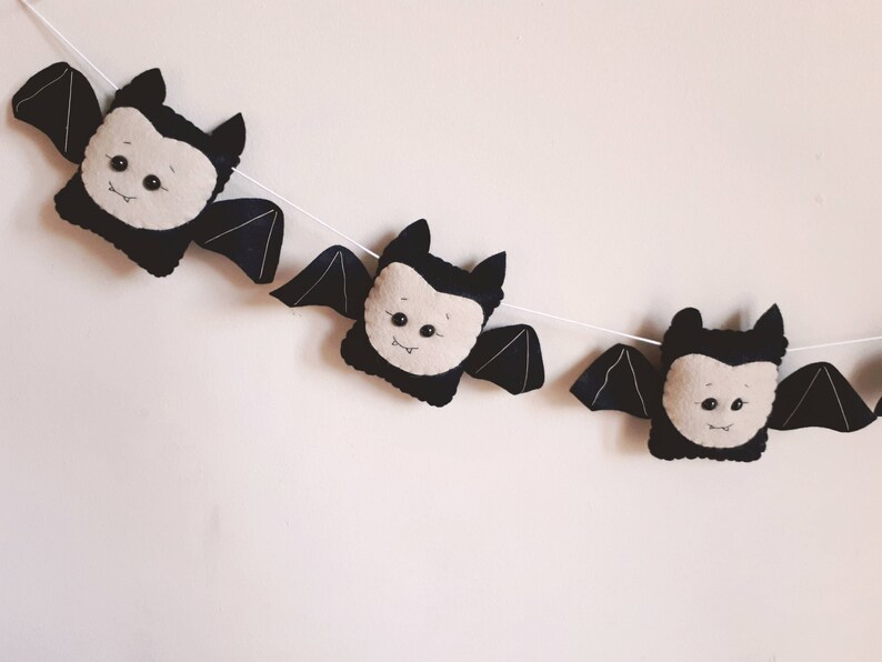 May include: A decorative garland featuring three plush bats. Each bat has black wings and body, with a cream-coloured face. The bats are strung on a white string, ideal for Halloween.
