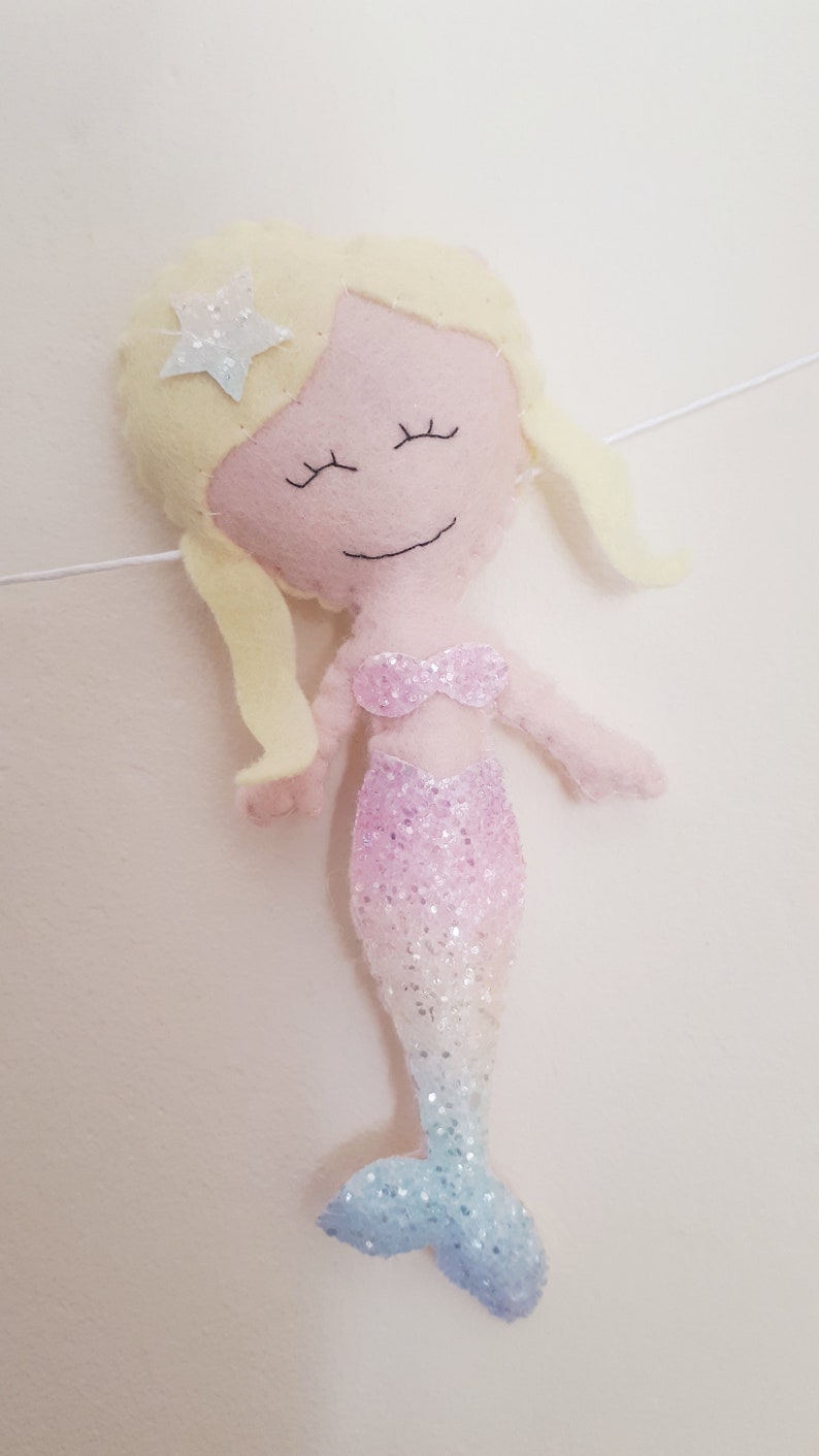 May include: A pink felt mermaid with a sparkly tail and blonde hair. The mermaid has a star on her head and closed eyes.