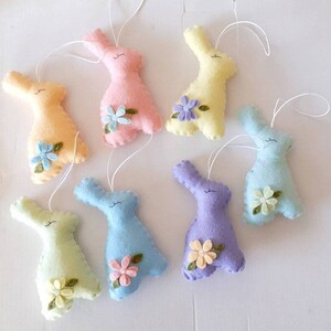 Peut inclure: Sept décorations de lapin en feutre de couleurs pastel, chacune avec une petite fleur en embellissement. Les lapins sont suspendus à une ficelle blanche.