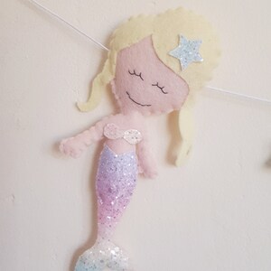 May include: A felt mermaid ornament with a pink face, blonde hair, and a sparkly purple and blue tail. The mermaid has a star on her head.