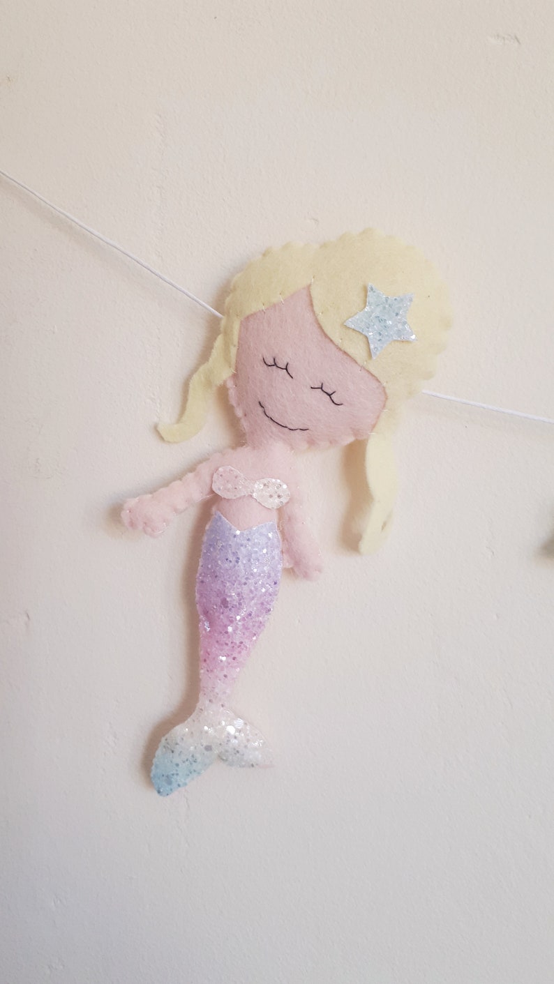 May include: A felt mermaid ornament with a pink face, blonde hair, and a sparkly purple and blue tail. The mermaid has a star on her head.