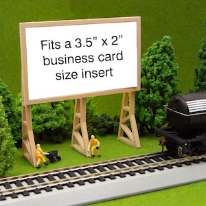 May include: A miniature model of a billboard with the text "Fits a 3.5" x 2" business card size insert" on a white background. The billboard is on a model train track with miniature trees and a model train car.