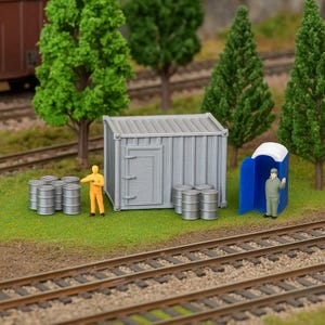 May include: Miniature gray corrugated metal storage container, barrels, and a blue portable toilet with a figure inside, set on a grassy area with model train tracks and trees. A figure in an orange jumpsuit stands nearby.