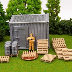 May include: A miniature model of a shipping container with a worker figure, barrels, and wooden pallets. The container is gray and has a door. The worker is wearing a yellow shirt and holding a tool. The barrels are silver and the pallets are brown.
