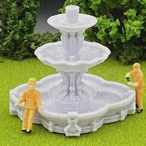 May include: A miniature white fountain with multiple tiers and ornate detailing. Two small figures in orange clothing are positioned near the fountain, suggesting a garden or landscape scene. The fountain is set on a green surface with green foliage in the background.