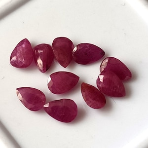 Puede incluir: Diez piedras preciosas de rubí en forma de pera, cada una con un tono rojo intenso y un corte facetado. Las piedras preciosas están dispuestas sobre una superficie blanca.