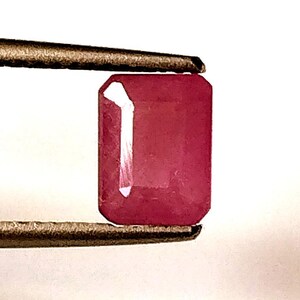 May include: A vibrant pink gemstone with a rectangular emerald cut. The gemstone is held in a pair of tweezers.