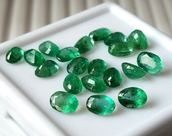 May include: A collection of oval-shaped, faceted emerald gemstones in various sizes, displayed on a white surface.