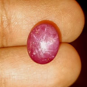 Peut inclure: Une pierre précieuse de forme ovale taillée en cabochon, d'une couleur rouge magenta profond. La surface de la pierre présente une texture fibreuse et chatoyante visible, avec une lumière se reflétant au centre, créant un effet étoilé subtil.