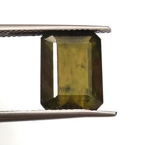 以下が含まれることがあります： ピンセットで挟まれた、長方形のオリーブグリーンの宝石。 宝石はファセットカットで、わずかに半透明に見えます。 無地の白い背景が宝石の色と形を際立たせています。