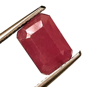 May include: A deep red emerald cut gemstone held by a pair of tweezers.