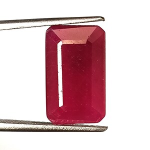 May include: A rectangular, faceted ruby gemstone held by tweezers. The ruby is a deep, rich red colour with a polished surface. The gemstone's shape is an emerald cut, with a flat top and bottom and bevelled sides.
