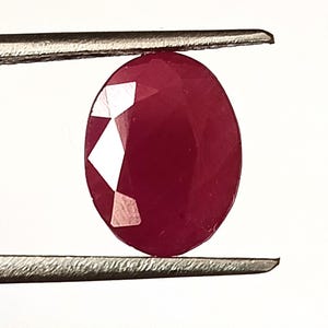 May include: A faceted, oval-shaped ruby gemstone held by tweezers. The ruby is a deep, rich red color with multiple facets that reflect light. The gemstone is set against a white background.