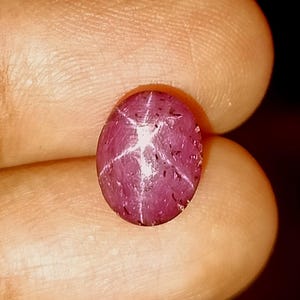 Op de afbeelding: Een ovaalvormige stersaffier edelsteen in cabochon-slijpvorm met een diepe magenta-rode tint. De steen vertoont een duidelijk zesstralig stereffect, bekend als asterisme, gecentreerd op het gepolijste oppervlak, samen met zichtbare natuurlijke insluitingen.