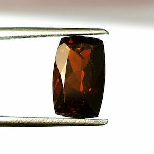 May include: A cushion-cut gemstone with a deep reddish-brown color and a faceted surface. The stone is held by a pair of silver-toned metal tweezers, displaying its translucent quality and geometric shape against a plain, light-colored background.